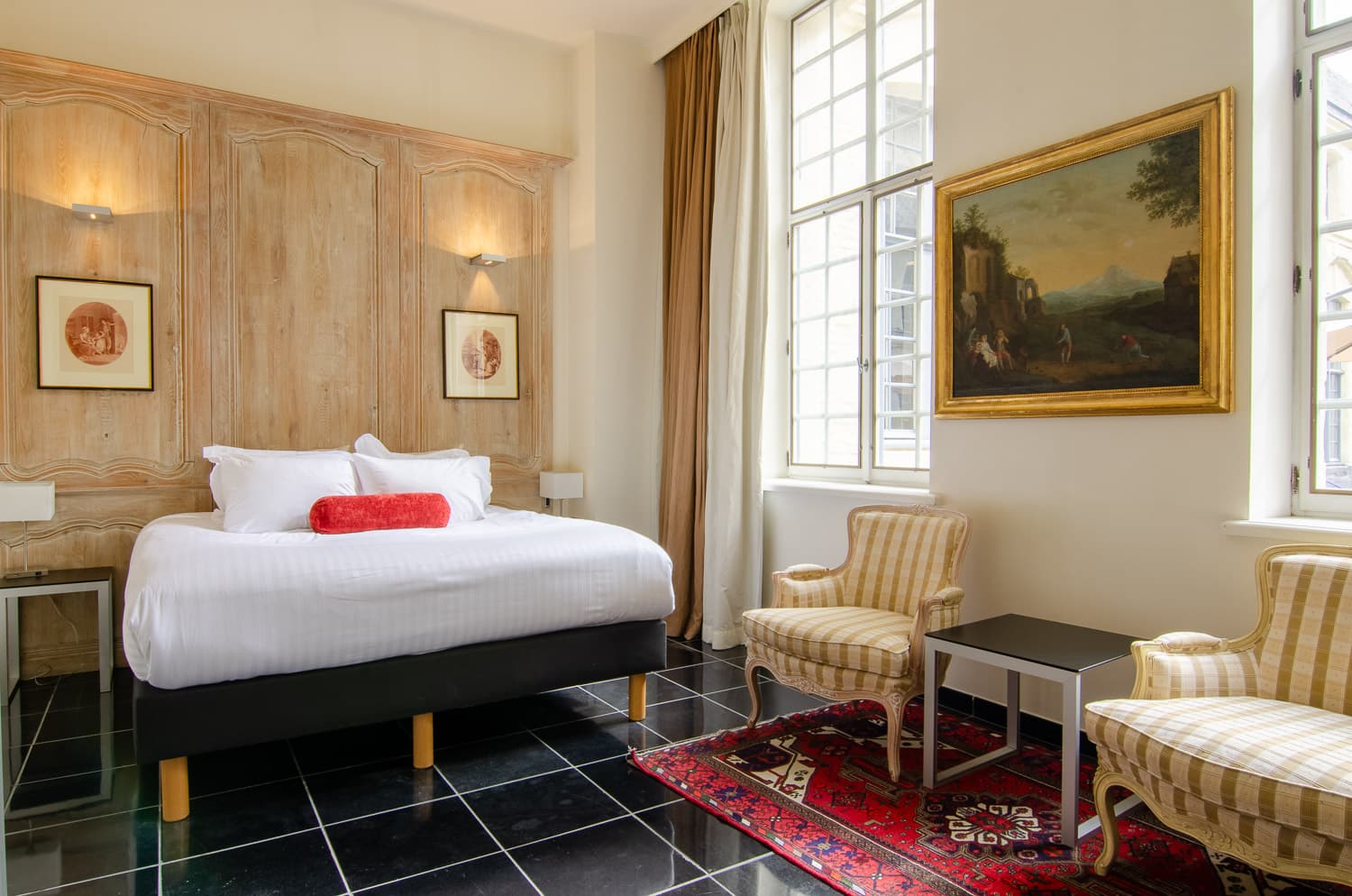 chambre d'hôtel classique avec lit à coussin rouge, fauteuils à carreaux et tapis oriental